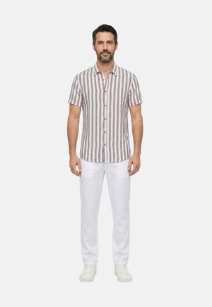 Uomo con capelli scuri e barba che indossa una camicia a maniche corte marrone e bianca a righe, pantaloni bianchi e scarpe da ginnastica bianche, che sta in piedi rivolto in avanti.