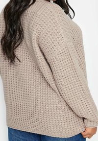 Kvinna i en beige grovstickad tröja och blå jeans, som visar sid- och baksidan av tröjans strukturerade mönster.