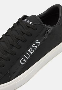 Black sneakers made of synthetic material featuring a textured upper, round toe, flat white rubber sole, and side zip detail with logo.