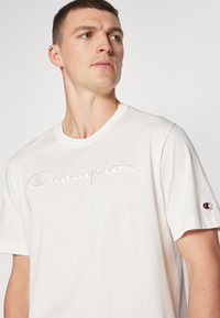 Weißes Baumwoll-T-Shirt mit gesticktem "Champion"-Logo in heller Fadenfarbe, rundem Ausschnitt und kurzen Ärmeln. Mit kleinem Champion-Logo am linken Ärmel.