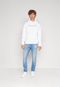 Fehér kapucnis pulóver fekete 'TOMMY JEANS' logóval, világoskék farmernadrággal és fehér sportcipővel párosítva, minimalista dizájnnal készült teljes szett.