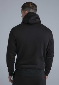 Uomo tatuato con indosso una felpa nera con cappuccio, visto di spalle su uno sfondo grigio uniforme.