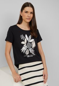 Zwart t-shirt met korte mouwen met witte en grijze bladerenprints en tekst. Zacht materiaal, relaxte pasvorm, ronde halslijn. Draagt met een gestreepte rok.