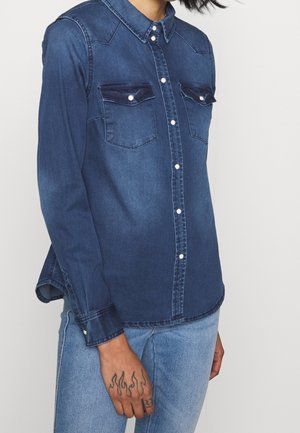 Person iført en mørkeblå denimskjorte med lange ærmer og knappede brystlommer samt lyseblå jeans, der viser en håndtatovering med flammedesign.