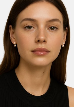 Deux boucles d'oreilles en perles avec une finition lisse et brillante ; une plus grande et une plus petite, montées sur une tige en ton or. Modèle portant un haut noir côtelé.
