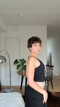 Mlada oseba s kratkimi kodrastimi lasmi nosi črn brezrokavnik in hlače ter stoji bok ob bok v svetlem, sodobnem bivalnem prostoru.