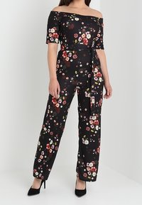 Combinaison noire à fleurs décolletée avec des motifs colorés, taille ceinturée, jambes larges et manches courtes ; associée à des escarpins noirs.