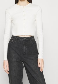 White long-sleeve cropped cardigan with a round neckline and pearl buttons. Paired with high-waisted black denim jeans.