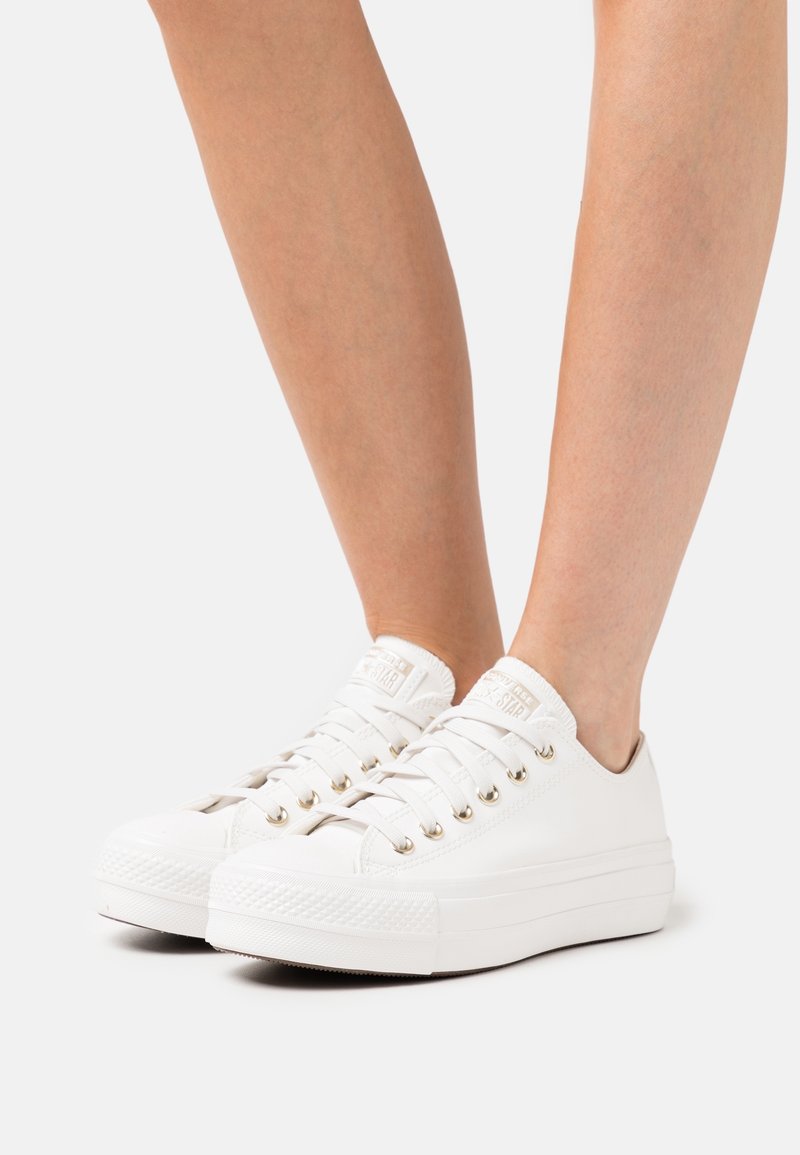 Weiße Low-Top-Sneaker mit Metallösen, die barfuß vor weißem Hintergrund getragen werden.