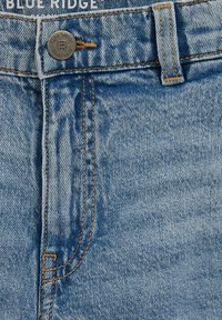 Heledat sinise teksapüksid, millel on metallist nööbi kinnis, oranž õmblemine ja kergelt tuhmuv muster, keskendudes esi tasku ja vöökoha piirkonnale.