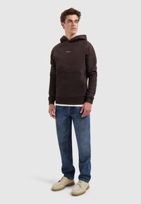Jonge man in een donkerbruine hoodie met de tekst "PURE PATH", blauwe spijkerbroek en beige veterschoenen, staand met ontspannen handen langs zijn zij.