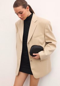 Bež oversize blazer z gumbom in enim žepom na prsih, nosi se nad črno oprijeto obleko, drži črno torbico. Gladka tekstura.