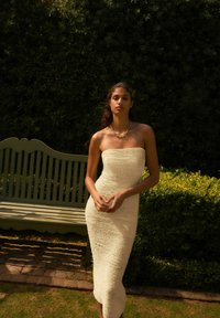 Robe midi sans bretelles en tissu crémé texturé, silhouette ajustée, avec des lignes verticales distinctes. Collier en chaîne argenté, banc vert et arrière-plan de feuillage.