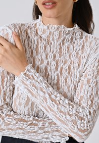 Top bianco in pizzo con un motivo testurizzato, scollo alto e polsini arricciati. Materiale trasparente con dettagli intricati e una forma aderente.