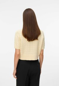Crème-kleurig, kort mouwend kabelgebreid truitje met een cropped ontwerp. Glad textuur, met een ribgebreide zoom en ontspannen pasvorm. Gecombineerd met zwarte broeken.