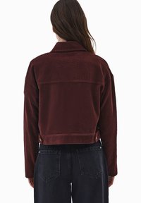 Burgundowa kurtka z kordurowego materiału z kołnierzem, skróconym krojem i widocznymi detalami szwów. Połączona z ciemnymi dżinsami, prezentując tylny widok.