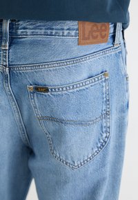 Jean en denim bleu clair avec des surpiqûres contrastées, présentant une patch en cuir marron avec le logo "Lee" et une poche arrière unique avec une étiquette.