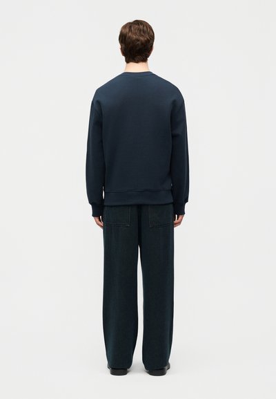 Άνδρας που στέκεται πλάτη, φορώντας σκούρο μπλε φούτερ και φαρδιά μαύρα τζιν με φαρδιά παντελόνια, μπροστά σε μια απλή ανοιχτόχρωμη επιφάνεια.