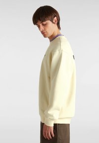 Lichtgele sweatshirt met een ronde hals en lange mouwen, met een subtiele logo op de voorkant en geribbelde manchetten, gepresenteerd aan de zijkant van een model.