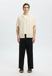 Camicia beige a maniche corte con dettagli in pizzo, abbinata a pantaloni neri a gamba larga e sandali marroni. Texture semplice e vestibilità rilassata.