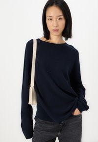 Mørkeblå, langermet genser med en avslappet passform og bred utringning; brukt sammen med mørkegrå jeans og en lys farged crossover-veske.