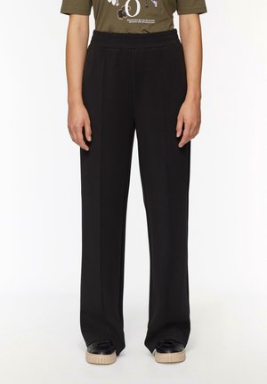 Osoba ubrana w luźne czarne spodnie z szwami z przodu, oliwkowo zielony t-shirt z nadrukiem oraz czarne trampki z beżowymi podeszwami, stojąca na białym tle.