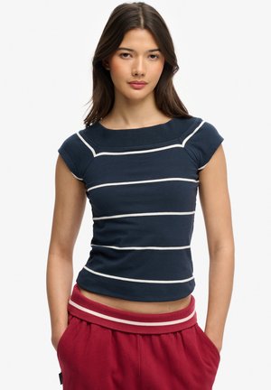 Jonge vrouw met lang donker haar, gekleed in een marineblauw gestreept topje met korte mouwtjes en rode broek, met handen in de zakken.