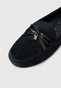 Σκούρο πράσινο μοκασίνι από σουέντ με ραμμένο σχέδιο, επίπεδη καουτσούκ σόλα, δερμάτινο φιόγκο και μικρή χρυσή λεπτομέρεια κλειδαριάς.