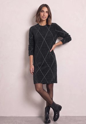Une femme porte une robe en maille couleur anthracite à motif losanges, des collants transparents et des bottines noires sur un fond de studio clair.