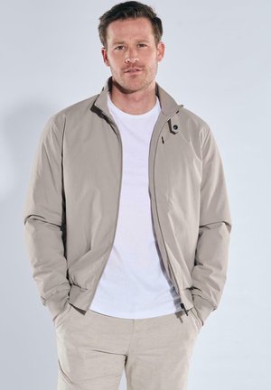 Man wearing light beige zip-up jacket over white t-shirt and beige pants, hands in pockets, standing against plain light background.