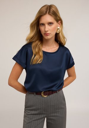 Blonde vrouw in een marineblauwe satijnen blouse met korte mouwen, grijs gestreepte broek, bordeauxrode riem en grote gouden creolen, staande met de handen achter de rug.