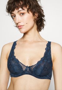 Donkerblauwe kanten beha met gekartelde randen en een doorschijnende mesh middenstuk. Bevat een klein decoratief accent in het midden en brede banden.