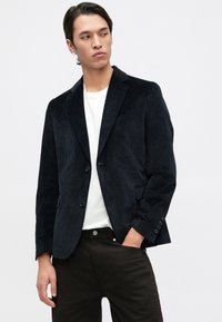 Blazer de veludo cotelê escuro com dois botões, lapelas com entalhe e bolsos laterais. Usado sobre uma t-shirt branca, combinado com calças escuras.
