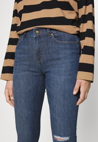 Jeans de ganga azul de cintura alta com uma textura suave, apresentando um fecho de botão dourado e um detalhe de joelho desgastado.