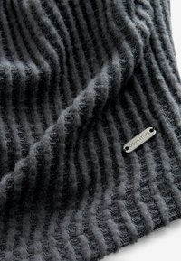 Strickschal in Dunkelgrau mit vertikaler Rippenstruktur und weichem, flauschigem Finish. Mit einem kleinen Metallschild, das mit "DOWNTIME" graviert ist.