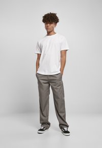 White crew-neck t-shirt, gray wide-leg trousers, black-and-white sneakers. Model stands with hands in pockets, short curly hair.