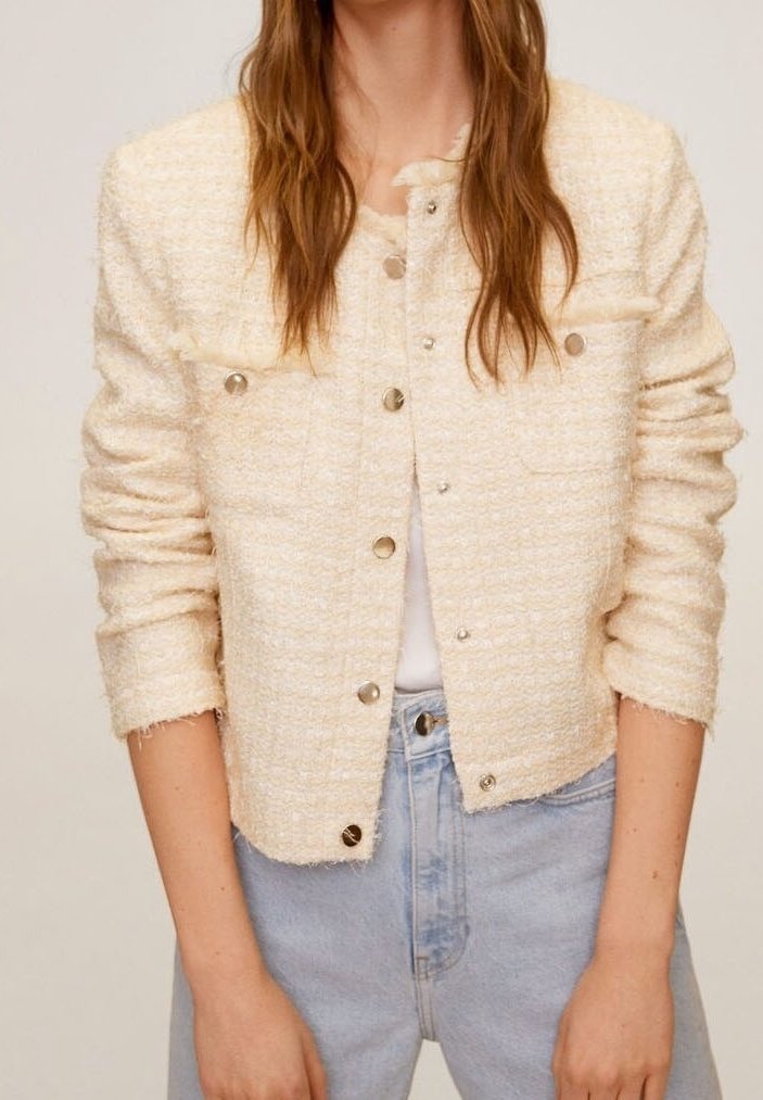 Femme portant une veste crème texturée boutonnée à manches longues et un jean taille haute bleu clair, debout devant un fond uni.