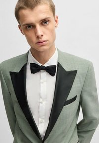 Giacca da smoking verde chiaro con risvolti in satin nero, abbinata a una camicia bianca e un papillon nero. Presenta una tasca singola a sinistra.