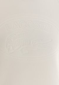 Camisa de algodón blanca con un logo de Lacoste en relieve y diseño de cocodrilo, con una textura acanalada y un acabado suave.