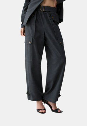 Donna che indossa pantaloni grigio scuro a vita alta e gamba larga con tasche cargo e tacchi alti neri open toe su sfondo bianco.
