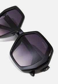 Ochelari de soare hexagonali cu un cadru din plastic lucios de culoare neagră și lentile gradient în nuanțe de violet. Prezintă o balama din metal cu un mic detaliu decorativ.