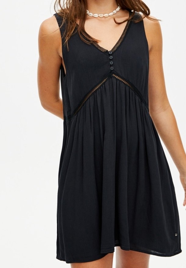 Robe noire sans manches avec col en V, trois boutons sur le devant, détails en dentelle et jupe ample plissée portée par une personne avec un collier en coquillage.