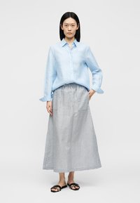 Femme portant une chemise bleu clair à manches longues boutonnée, une jupe rayée longue jusqu'aux chevilles avec taille à cordon et des sandales noires à enfiler.