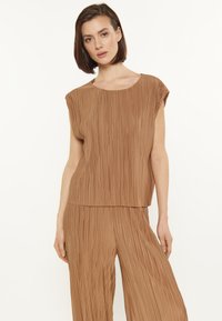 Top brown pliat cu guler rotund și mâneci scurte, fabricat dintr-un material moale; purtat împreună cu pantaloni largi pliati asortați.