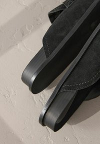 Sandales en suède noir avec une semelle en caoutchouc plate et des sangles larges. Les bords sont ornés de coutures, ajoutant une texture au design.