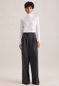 Kobieta o krótkich kręconych włosach nosi biały golf i szerokie, ciemnoszare spodnie z wysokim stanem, stojąc na jednolitym tle.