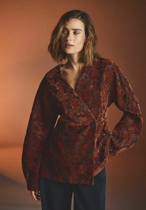 Femme aux cheveux mi-longs portant une veste cache-cœur texturée à motifs floraux rouge foncé et un pantalon foncé sur un fond orange atténué.