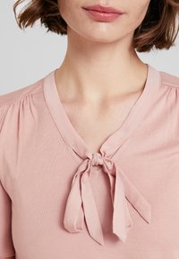 Jasnoróżowa bluzka z dekoltem w kształcie litery V i marszczeniem na ramionach. Z dodatkiem kokardki wiązanej przy szyi. Gładka tekstura materiału.