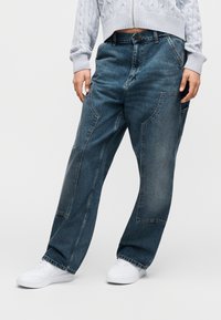 Άτομο που φοράει ανοιχτό μπλε κοντό πουλόβερ, σκούρα μπλε άνετο τζιν με μεγάλες τσέπες και λευκά αθλητικά παπούτσια, στέκεται μπροστά σε απλό φόντο.