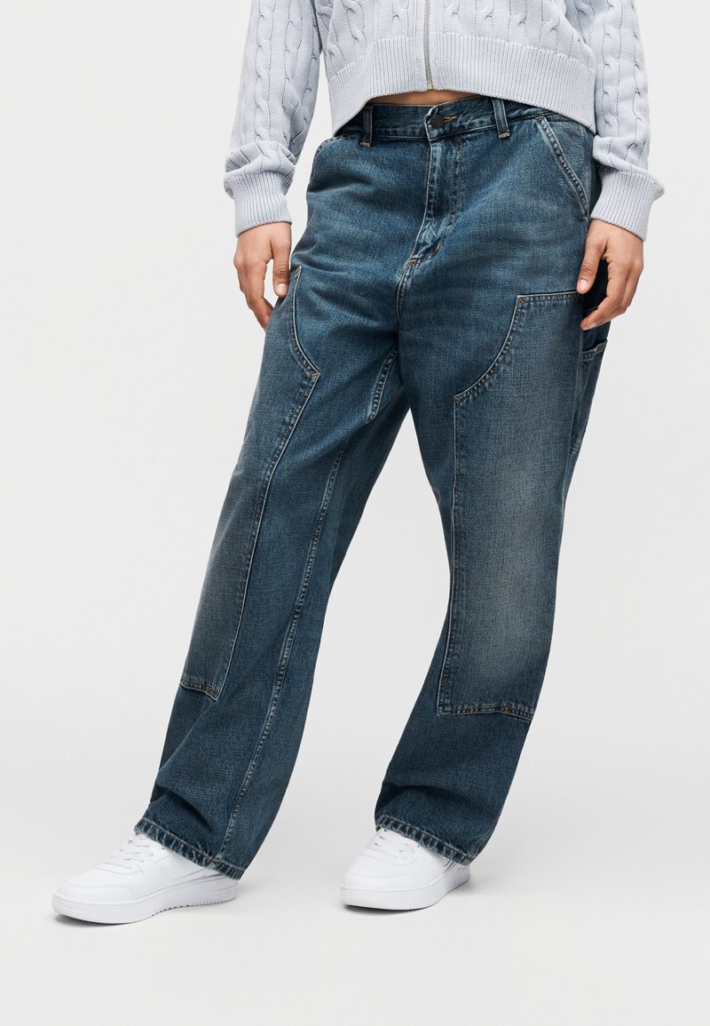 Personne portant un pull court bleu clair, un jean ample bleu foncé avec de grandes poches, et des baskets blanches, debout devant un fond uni.